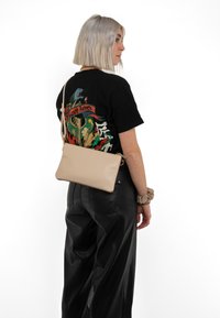 Beige bőrből készült keresztpántos táska sima textúrával, téglalap alakú, arany tónusú cipzárral. A modellen grafikus fekete póló és bőrnadrág található.