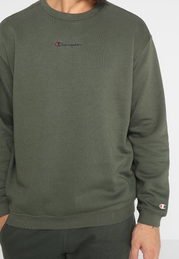 Homme portant un sweat-shirt Champion vert olive avec logo sur la poitrine et la manche, debout sur un fond blanc uni.