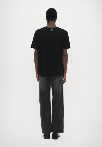 Black cotton t-shirt with short sleeves, straight cut, and logo detail at the back, paired with loose-fitting dark grey jeans and black shoes.