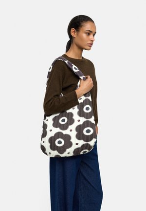 Kobieta o ciemnych włosach zaczesanych do tyłu, ubrana w brązowy sweter i niebieskie spodnie, niesie dużą białą torbę z czarnymi i czerwonymi wzorami kwiatowymi.