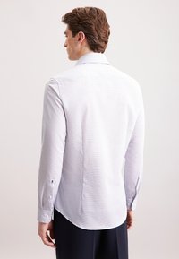 Uomo con capelli castani che indossa una camicia bianca a maniche lunghe aderente e pantaloni neri, visto di spalle su uno sfondo semplice.