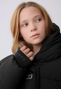 Zwarte gewatteerde jas met een hoge kraag en elastische manchetten. Heeft een gestructureerd oppervlak en een zilverkleurig logo op de borst.