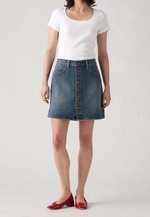 Femme portant un haut blanc à manches courtes, une jupe en jean bleue avec des boutons sur le devant et des chaussures rouges à talons bas, debout devant un fond uni.