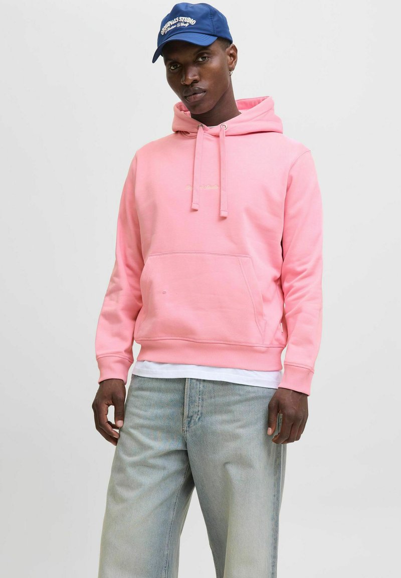 Felpa rosa con cappuccio, dotata di tasca frontale e coulisse. Indossata sopra una maglietta bianca, abbinata a pantaloni in denim azzurri chiari.