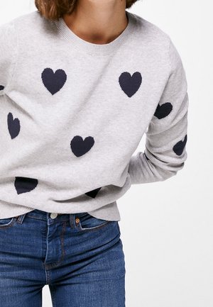 Vrouw in een lichtgrijze sweater met zwarte hartpatronen, gecombineerd met blauwe denim jeans, tegen een effen witte achtergrond.