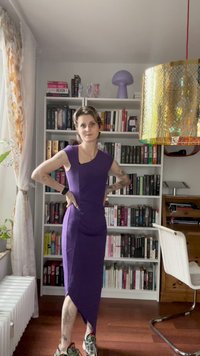 Violetas spalvos prigludusi suknelė su trumpomis rankovėmis ir asimetriška pakraščiu. Lygus audinys, iki kelių, derinama su raštuotais sportbačiais. Fone knygų lentyna.