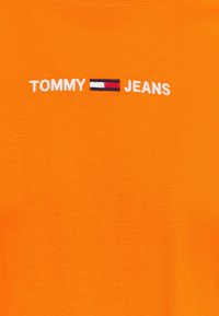 Pomarańczowa bawełniana koszulka z białym haftem tekstu "TOMMY" nad wzorem flagi i "JEANS" poniżej. Klasyczny okrągły dekolt, gładka faktura.