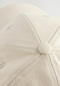 Gorra de béisbol color beige fabricada en tela texturizada, con corona redondeada, paneles cosidos y un botón circular en la parte superior.