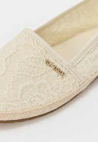 Beige slip-on kengät, joissa on pitsi päällyste, kultasävyinen "Tom Tailor" -logo ja kuvioitu, kudottu juuttipohja.