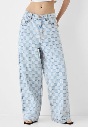 Lichtblauwe wijde spijkerbroek met ruitpatroon, gedragen met zwarte sandalen met open teen en een cropped wit topje.