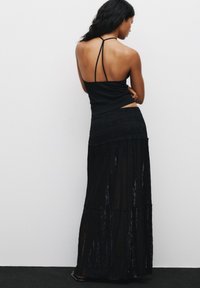 Haut à col dégagé noir avec des fines bretelles, associé à une longue jupe noire superposée avec un tissu transparent et texturé et des accents horizontaux.