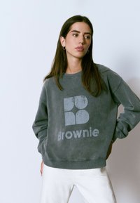 Sudadera gris de cuello redondo con un logo desgastado que dice "Brownie" en el frente. Tejido suave con puños y dobladillo acanalados. El modelo lleva pantalones blancos.