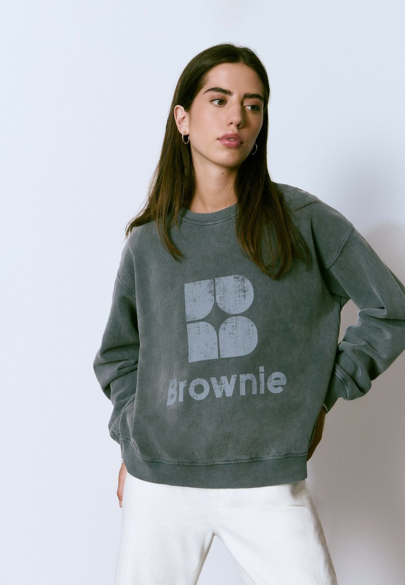 Sudadera gris de cuello redondo con un logo desgastado que dice "Brownie" en el frente. Tejido suave con puños y dobladillo acanalados. El modelo lleva pantalones blancos.