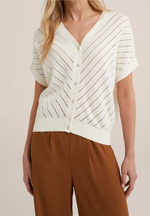 Femme portant un cardigan en maille blanc à manches courtes, boutonné, avec des trous diagonaux, et un pantalon marron taille haute à jambes larges.