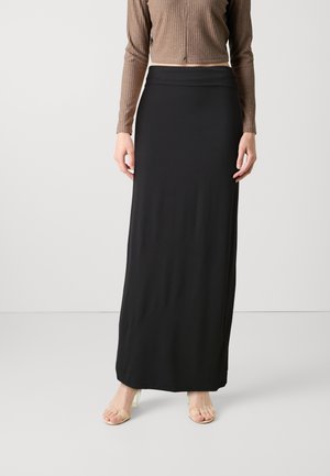 Žena oblečená v dlouhé černé sukni s vysokým pasem, hnědém žebrovaném topu s dlouhým rukávem a sandálech na podpatku s průhledným páskem, stojící na bílém pozadí.