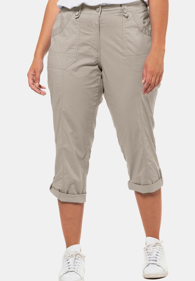 Pantalon beige court avec revers roulés porté avec des baskets blanches, montrant les poches avant et les détails de couture.