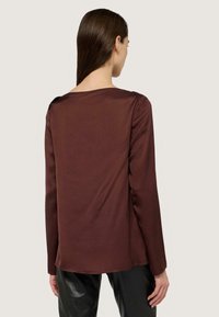 Maglia marrone a maniche lunghe, texture liscia, scollatura arrotondata, arricciature sulle spalle e orlo posteriore allungato, abbinata a pantaloni neri lucidi.