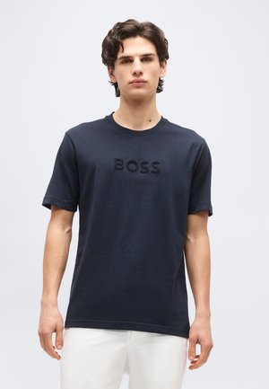 Νεαρός άντρας με σκούρα μαλλιά που φοράει μαύρο T-shirt με το κείμενο «BOSS» και λευκό παντελόνι, στέκεται μπροστά σε απλό φωτεινό φόντο.