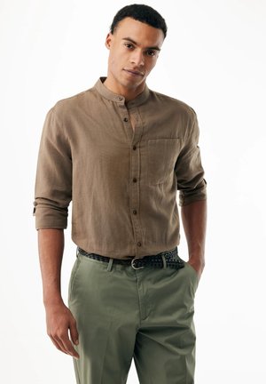 Uomo con capelli corti e ricci che indossa una camicia marrone con collo alla coreana e pantaloni verdi con una cintura intrecciata nera, mani nelle tasche.