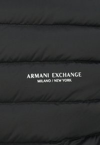 Sort polstret jakke med horisontal quiltning. Har hvid "Armani Exchange MILANO / NEW YORK" logo på fronten. Glat, mat overflade.