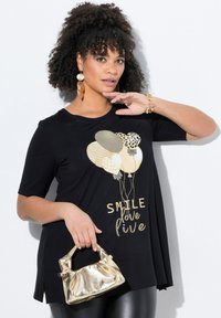Černé tričko s krátkým rukávem a zlatou grafikou balónku a textem "SMILE love live." Modelka drží lesklou zlatou kabelku.