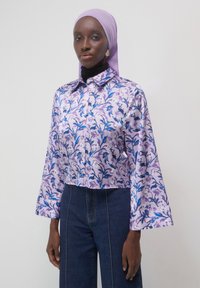Frau in einer blasslila blumigen Bluse mit weiten Ärmeln, dunkelblauen hoch taillierten Jeans und einem passenden blasslila Kopftuch.