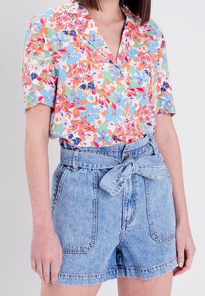 Femme portant un chemisier à manches courtes à motif floral dans des couleurs pastel et un short en jean bleu clair taille haute noué avec une ceinture.