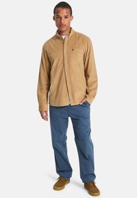 Κοντουνάρι από ταμπά κορδούρα με τσέπη στο στήθος, συνδυασμένο με μπλε παντελόνι και καφέ σουέντ παπούτσια, στέκεται μπροστά σε λευκό φόντο.