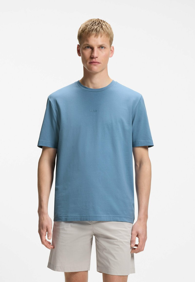 Jeune homme aux cheveux blonds courts portant un t-shirt BOSS bleu clair et un short beige, debout devant un fond blanc.