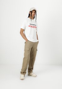 Balta T-kreklu ar uzrakstu "new balance ATHLETICS", khaki krāsas cargo bikses, bēšas sporta kurpes un gaišs cepure. Vienkāršs dizains, brīvs fit.