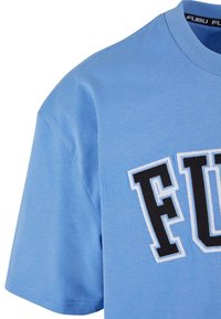 Tricou scurt cu mânecă scurtă, de culoare albastru deschis, confecționat dintr-un material moale, având un logo brodat „FUBU” în alb și negru, afișat proeminent pe față.