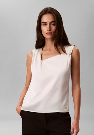 Femme aux longs cheveux bruns portant un haut blanc sans manches et un pantalon noir, debout devant un fond gris uni.