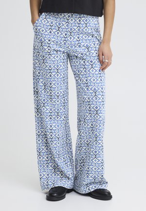 Personne portant un pantalon large à motif géométrique bleu et blanc, des mocassins noirs, une main dans la poche, debout devant un fond uni.