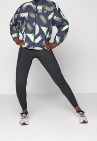 Kurtka przeciwwietrzna New Balance w kolorze granatowym z abstrakcyjnymi zielonymi i beżowymi wzorami, zestawiona z czarnymi legginsami i białymi trampkami z różowymi akcentami.