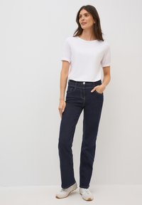 Chemise blanche à manches courtes associée à un jean droit bleu foncé. Les jeans comportent des poches avant et des coutures dorées. Portés avec des baskets claires.