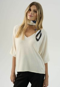 Krémový sveter s výstrihom do V a širokými rukávmi, vyrobený z ľahkého materiálu. Obsahuje malú zlatú emblematickú značku na leme, nosený s vzorovaným šálom.