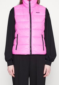 Person trägt eine leuchtend pinkfarbene gesteppte Weste mit schwarzem Reißverschluss und Taschen über einem langärmligen schwarzen Oberteil und schwarzen Hosen.