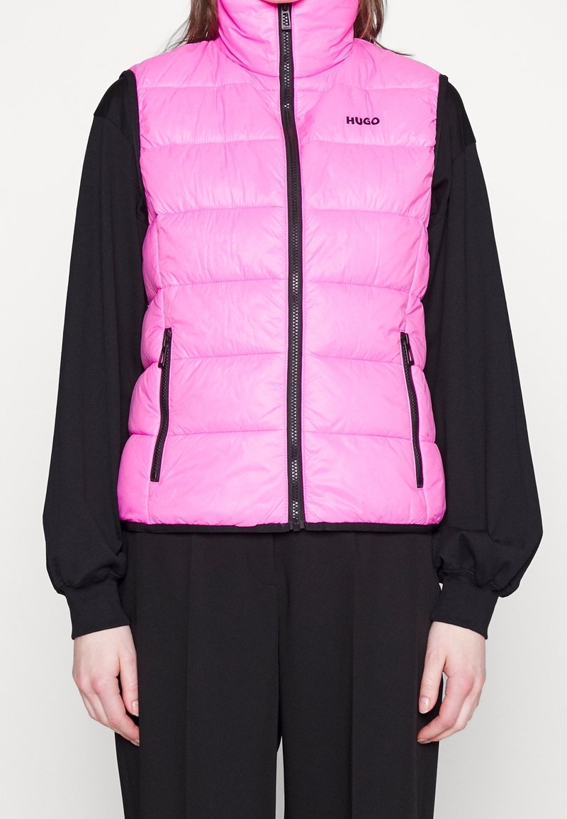 Person trägt eine leuchtend pinkfarbene gesteppte Weste mit schwarzem Reißverschluss und Taschen über einem langärmligen schwarzen Oberteil und schwarzen Hosen.