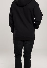 Personne debout, de dos, portant un sweat à capuche noir et un jean noir, sur un fond clair et uni.