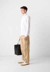 Hvit langermet skjorte med knapper, beige bukser og hvite sneakers med brune detaljer; bærer en svart totebag. Glatt stoff og avslappet passform.