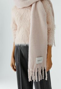 Helles rosa Wollschal mit fransigen Kanten, ausgestattet mit einem weißen Etikett mit dem Text "oui". Getragen über einem flauschig strukturierten rosa Pullover und gestreiften Hosen.