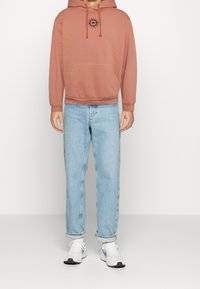 Hoodie di cotone color rosa con una grafica del sole sul petto, abbinato a jeans dritti blu chiaro e sneakers bianche.