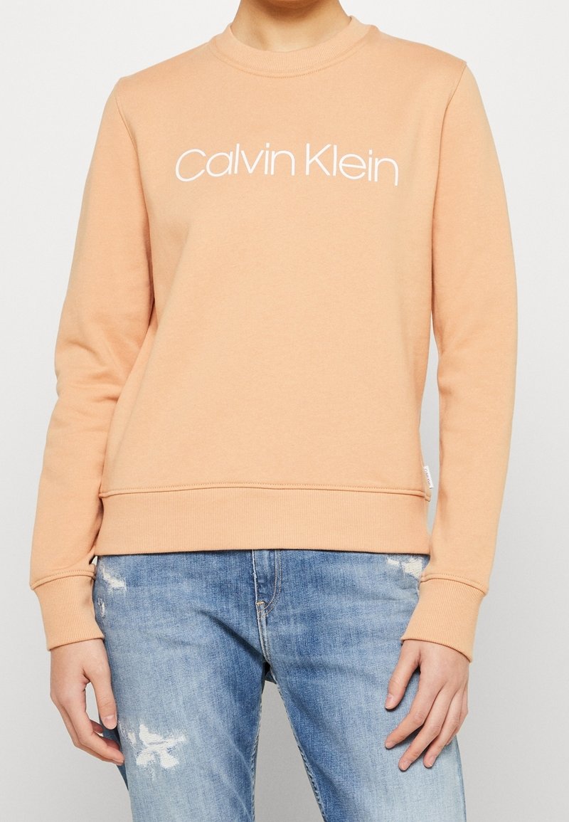 Personne portant un sweat-shirt Calvin Klein orange clair et un jean bleu avec des déchirements légers aux cuisses et aux genoux.