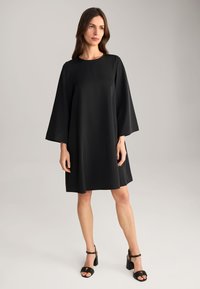 Zwarte A-lijn jurk met lange klokmouwen, ronde halslijn en gladde stof. Draagt een paar chunky zwarte haksandalen met een gouden accent.