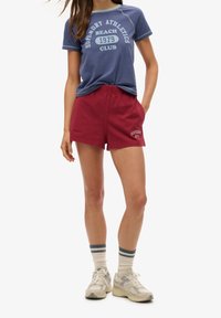 Camiseta de algodón azul con texto blanco, combinada con pantalones cortos burdeos con un logo bordado. El modelo también lleva zapatillas blancas con acentos azules.