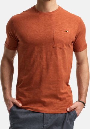 T-shirt à manches courtes couleur rouille fabriqué en tissu texturé, avec une poche avant ornée d'un petit logo. Design simple avec col rond.