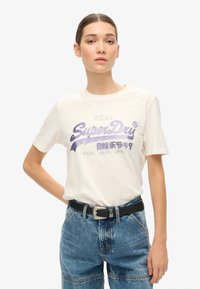 Cremefarvet kortærmet T-shirt med metallisk lilla "SuperDry" grafik, parret med blå højtaljede denimshorts og sort bælte.