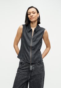 Juodas džinsinis liemenės liemenė be rankovių su priekiu užtrauktuku, smaigaliu apykakle ir prigludusiu dizainu. Dėvima su derančiomis aukšto liemens džinsinėmis kelnėmis.