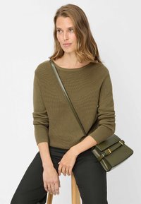 Olivgrüner, gerippter Pullover mit rundem Ausschnitt, kombiniert mit dunkler Hose. Eine Umhängetasche in passendem Grün mit goldfarbenen Beschlägen.