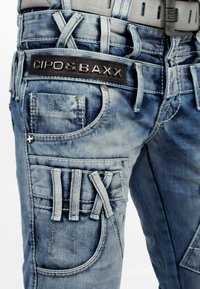 Pantalons cargo en denim bleu avec un patch logo noir, plusieurs poches, des coutures texturées et des détails de sangle uniques sur le côté.